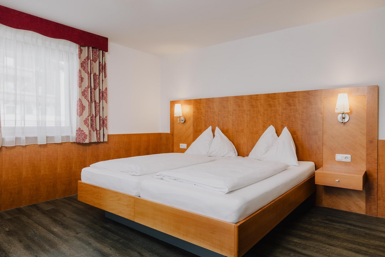Ein mit weißer Bettwäsche aufgedecktes Doppelbett mit hölzernem Kopfteil mit passenden Nachtkästchen. Links davon fällt Sonnenlicht durch ein Fenster.
