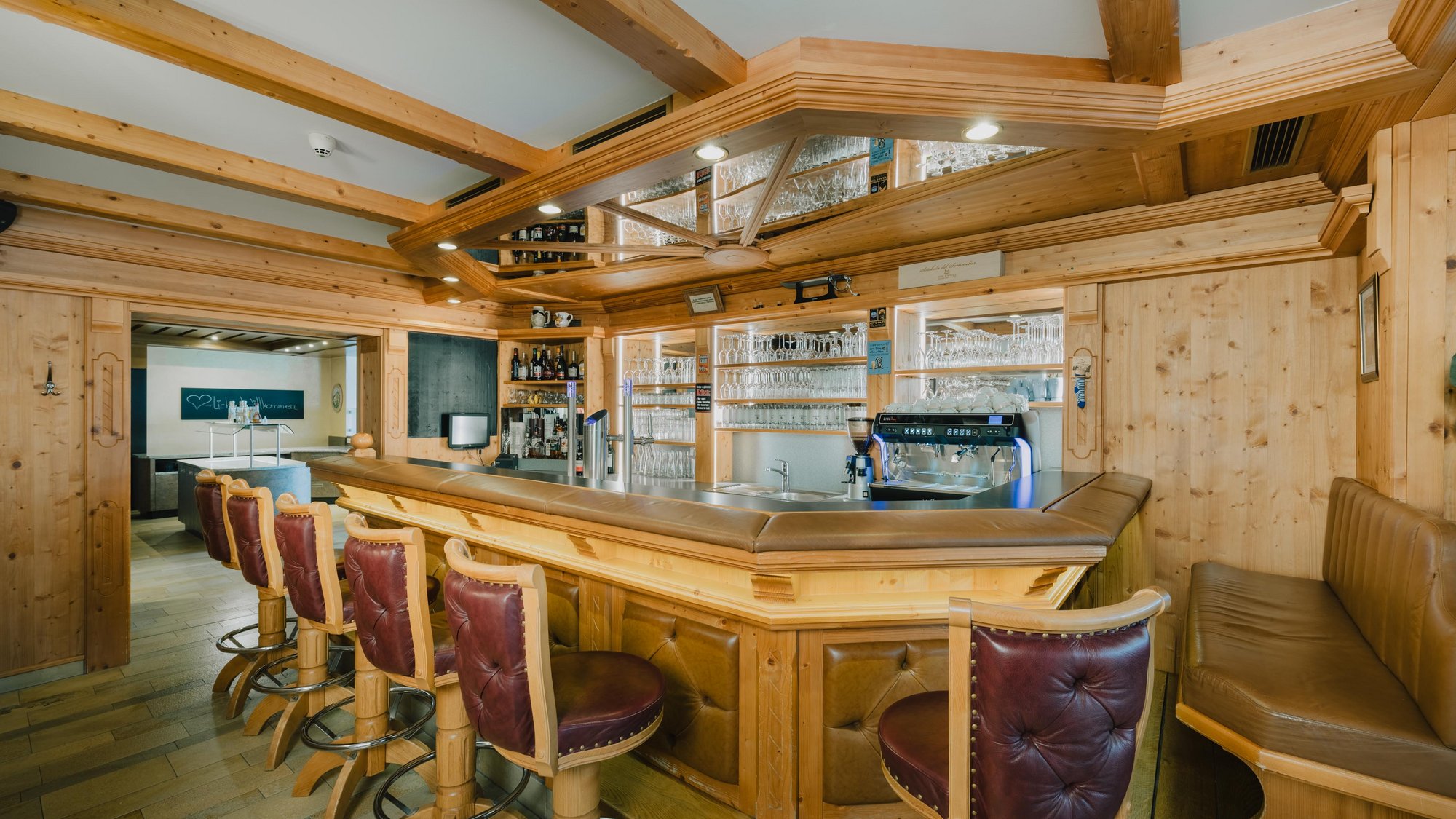 Eine Bar in einem holzvertäfelten Raum. Vor der Bar stehen Ledergepolsterte Barstühle, während im Hintergrund Regale mit verschiedenen Getränken und Gläsern sanft beleuchtet sind.