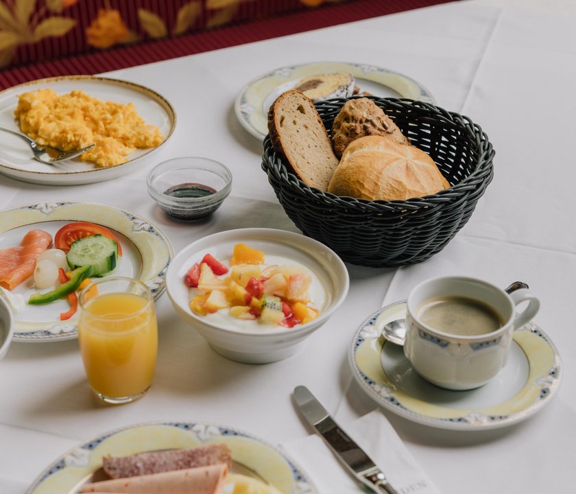 Ein reich gedeckter Frühstückstisch für zwei Personen. Auf einer weißen Tischdecke stehen ein Korb mit Brot, Joghurt mit Früchten, Rührei, Lachs, Orangensaft, Kaffee und einiges mehr.