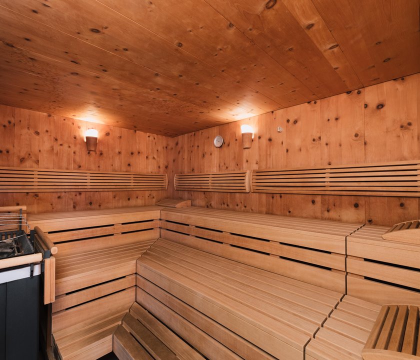 Blick in die Sauna. Komfortable Holzbänke liegen direkt gegenüber dem traditionellen Saunaofen. Warme Beleuchtung auf holzgetäfelten Wänden schafft eine gemütliche Atmosphäre.