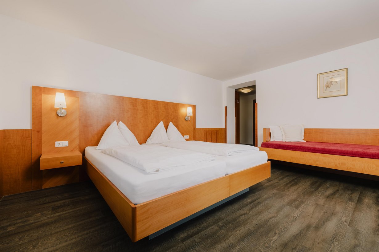 Ein mit weißer Bettwäsche aufgedecktes Doppelbett mit hölzernem Kopfteil mit passenden Nachtkästchen. Rechts davon führt ein Durchgang in den Vorraum.