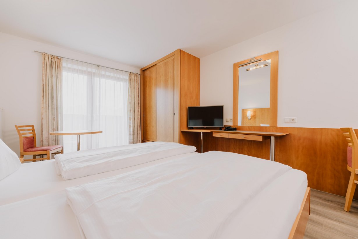 Blick über ein komfortables Bett in die gegenüberliegende Zimmerecke, die von einem Einbauschrank und einem kleinen Tisch mit Fernseher gefüllt ist. Links vom Bett steht ein Stuhl mit kleinem Tisch vor einer großen Balkontüre.