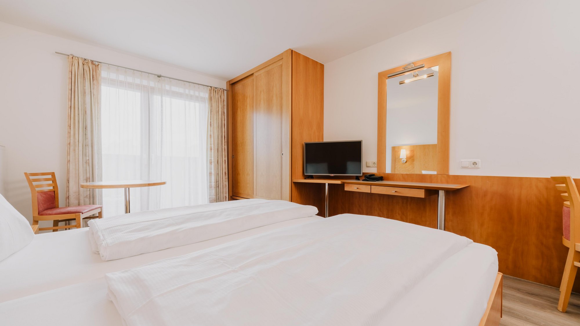   Blick über ein komfortables Bett in die gegenüberliegende Zimmerecke, die von einem Einbauschrank und einem kleinen Tisch mit Fernseher gefüllt ist. Links vom Bett steht ein Stuhl mit kleinem Tisch vor einer großen Balkontüre.