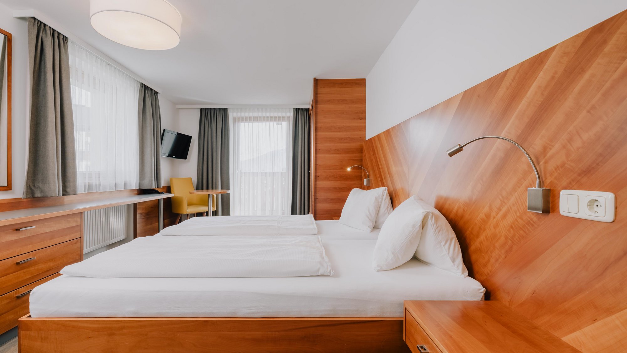 Ein komfortables Doppelbett mit hölzernem Kopfteil steht neben einem passenden Einbauschrank. Gegenüber dem Fußende befindet sich ein kleiner Schreibtisch. Zwischen einem gemütlichen Sessel und dem Einbauschrank ist fast die ganze Wand von einer Großen Balkontür mit weißen Gardinen ausgefüllt.
