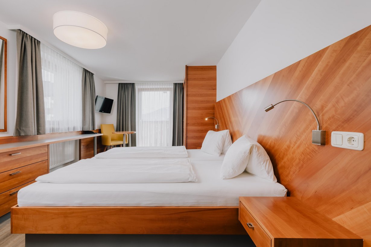 Ein komfortables Doppelbett mit hölzernem Kopfteil steht neben einem passenden Einbauschrank. Gegenüber dem Fußende befindet sich ein kleiner Schreibtisch. Zwischen einem gemütlichen Sessel und dem Einbauschrank ist fast die ganze Wand von einer Großen Balkontür mit weißen Gardinen ausgefüllt.