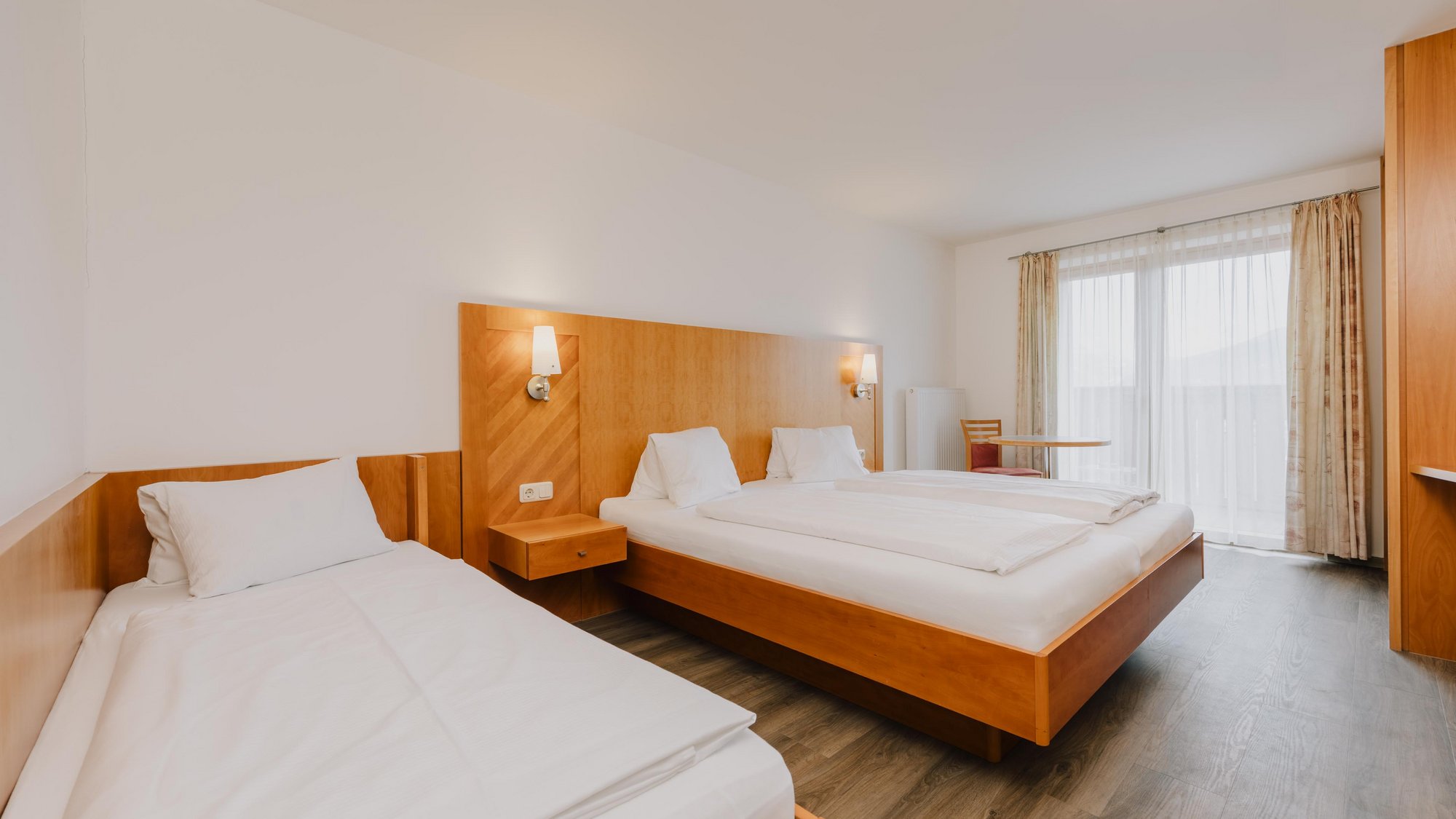 Neben einem mit weißer Bettwäsche aufgedecktes Doppelbett mit hölzernem Kopfteil steht ein weiteres Einzelbett. Der Raum wird durch eine große Balkontüre von Sonnenlicht erhellt.