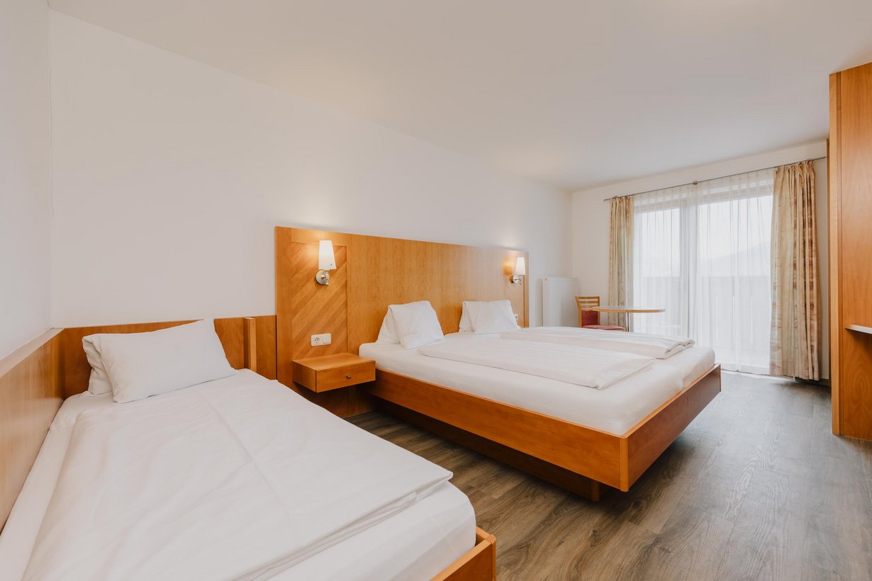 Neben einem mit weißer Bettwäsche aufgedecktes Doppelbett mit hölzernem Kopfteil steht ein weiteres Einzelbett. Der Raum wird durch eine große Balkontüre von Sonnenlicht erhellt.