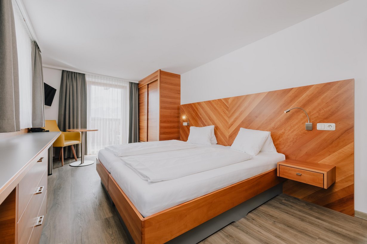 Ein komfortables Doppelbett mit hölzernem Kopfteil steht neben einem passenden Einbauschrank. Gegenüber des Fußendes befindet sich ein kleiner Schreibtisch und zwischen einem gemütlichen Sessel und dem Einbauschrank ist fast die ganze Wand von einer großen Balkontür mit weißen Gardinen ausgefüllt.