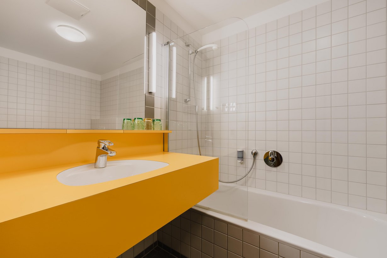 Modernes weiß gefliestes Badezimmer mit Badewanne und Glasduschwand. Ein großer beleuchteter Spiegel hängt über einem gelben Waschbeckentisch.