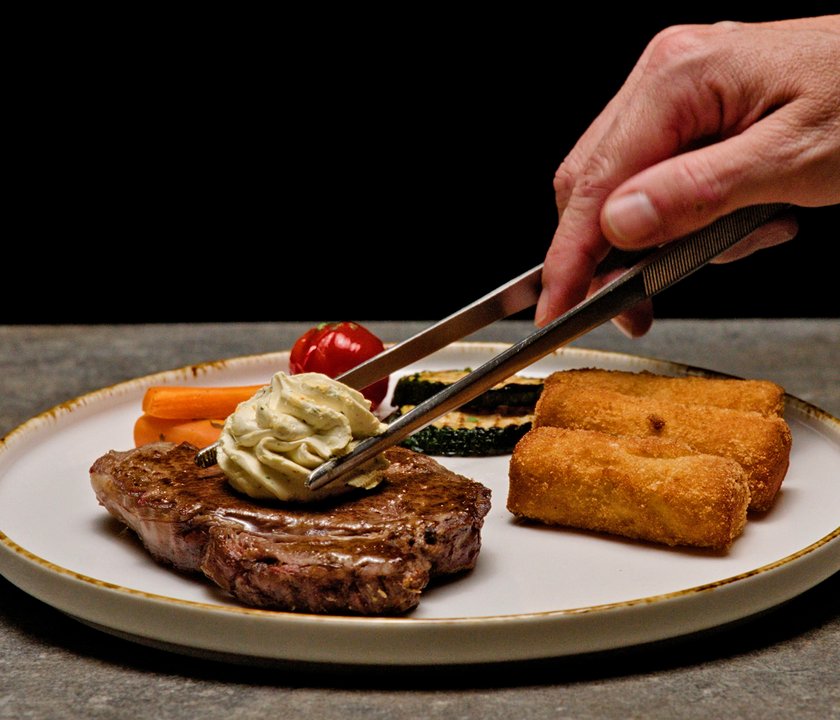 Auf einem weiten Keramikteller edel angerichtetes Steak mit Kroketten und gebratenem Gemüse. Eine Hand legt mit einer Zange ein Stück Kräuterbutter auf das Steak.