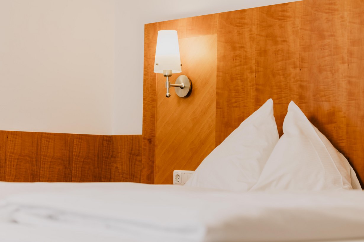 Neben einem Bett mit weißer Bettwäsche und hölzernem Kopfteil taucht eine kleine Lampe mit hellem Schirm den Raum in angenehmes Licht.