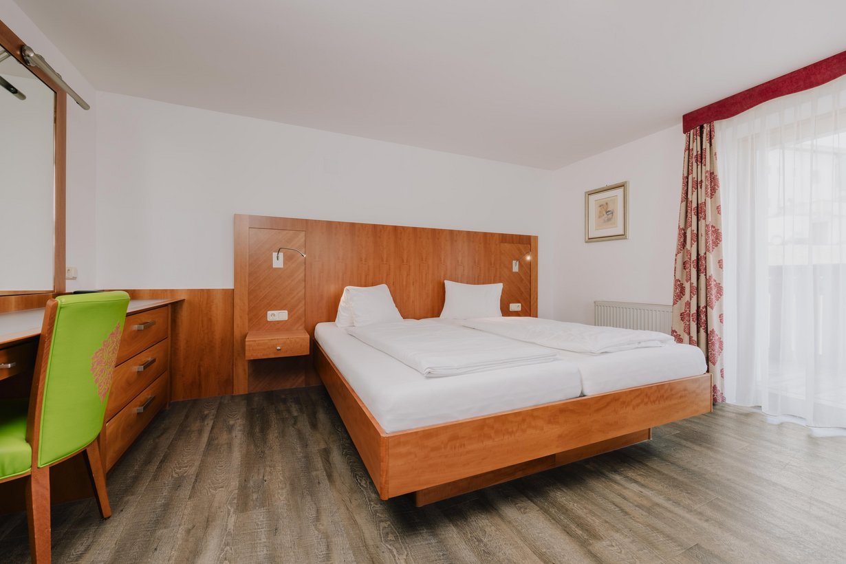 Neben einem komfortablen Doppelbett mit getäfelten Kopfteil steht an der linken Wand eine Hölzerne Kommode mit einem Schreibtisch und Stuhl daneben und Spiegel darüber.