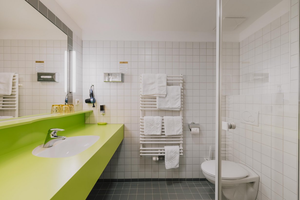 Modernes weiß gefliestes Badezimmer mit Duschkabine und beheiztem Handtuchhalter daneben. Ein großer beleuchteter Spiegel hängt über einem hellgrünen Waschbeckentisch gegenüber der Dusche.
